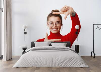 Portrait of young successful woman in glasses shows hand frame as if taking photo on camera and smiling, standing against white background Wall mural