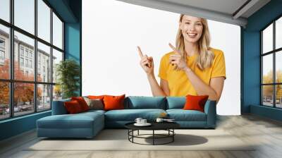 Portrait of young woman demonstrating, showing announcement or logo, pointing fingers left, standing in yellow t-shirt over white background Wall mural