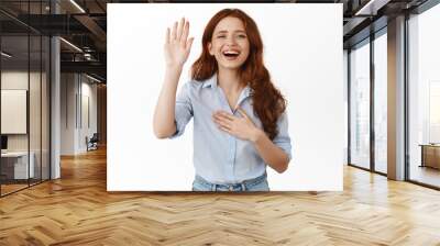 Smiling redhead girl introduce herself, raising hand and holding palm on heart, making promise, looking happy, tell her name, standing against white background Wall mural