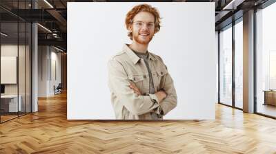 Studio shot fo confident friendly casual male redhead with beard and wavy hairstyle crossing hands on chest and smiling joyfully at camera gazing with satsfied and pleased look over gray wall Wall mural