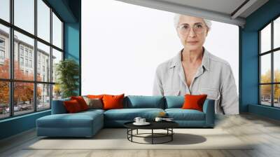 Studio shot of wise and intelligent skillful senior female doctotr in glasses with gray hair standing calm and confident against white background looking self-assured at camera Wall mural