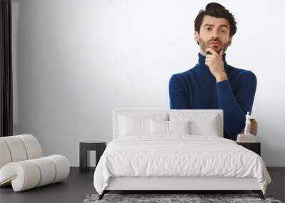 Stylish serious-looking young man with bristle in trendy high neck sweater, touching chin, rubbing beard and smirk as judging something, thinking or pondering checking out something interesting Wall mural