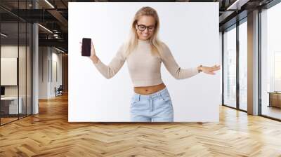 Technology and lifestyle concept. Carefree happy blond woman in glasses looking on floor as dancing carefree alone over white background listening music in wireless earbuds, holding smartphone Wall mural