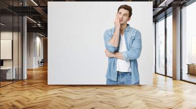 Tired and annoyed handsome boy holding palm on cheek and looking up, expressing boredom while standing over gray background. When you work time goes slow. Man in office counts minutes to go home Wall mural