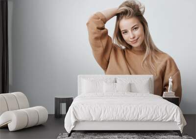 Young attractive woman wearing stylish long-sleeved sweatshirt looking with her dark appealing eyes at camera, smiling gently. Blonde caucasian female student posing against gray studio wall Wall mural