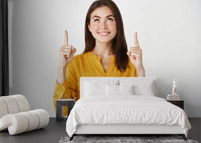 Young happy woman smiling, pointing and looking up at promo offer, concept of shopping and advertising, standing over white background Wall mural