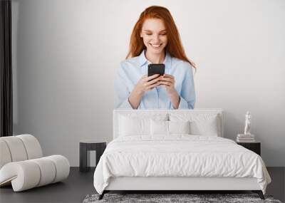 Young redhead woman reading message on mobile phone and smiling. Businesswoman looking happy at smartphone screen, standing over white background Wall mural