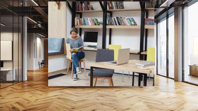 Young smart light-haired girl with bob hairstyle in casual clothes sitting on chair in modern library, reading favourite book, relaxing after long day on study Wall mural