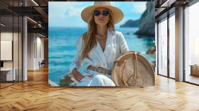 Outdoor summer fashion portrait of elegant woman wearing white linen suit, belt, straw hat, sunglasses, with round wicker bag, posing on rocks near sea. Copy, empty space for text illustration Wall mural