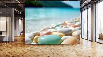 Vibrant, smooth pebbles of various colors lie on a scenic beach by the ocean. The sunny landscape and calm waters create a serene atmosphere. Wall mural