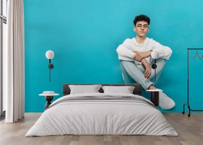 Confident teen boy in eyewear and white clothes, sitting down, looking at camera, isolated  turquoise background. Wall mural