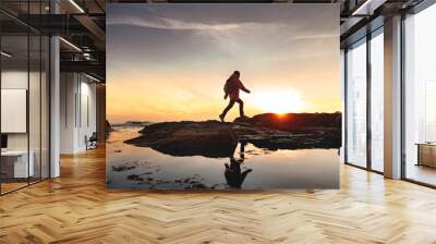 Female hiker silhouette with reflection in calm water is walking at seashore against sunset sky Wall mural