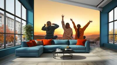 Three happy young girls are having fun and enjoys sunset from view point. Three silhouettes with open arms are sitting against sunset sky Wall mural