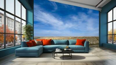 A bright blue sky with wispy white clouds above a dry badlands landscape.
 Wall mural