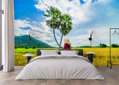 Vietnamese girl in dark red and bottle green traditional costume dress with conical hat walking on yellow green rice field and two palm trees, Ba Den mountain, blue sky in background. Wall mural