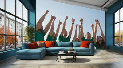 Group of happy young people in green T-shirts raise their hands up and show thumbs up, on the background of blue sky in yellow grass. Wall mural