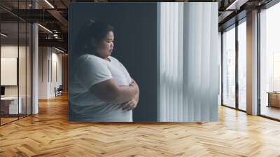 Lonely obese woman standing near the window Wall mural