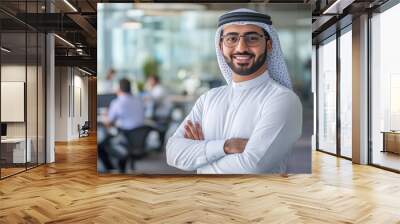 Portrait of a smiling Arabian man standing with arms crossed in a modern office, wearing casual business attire and glasses Wall mural