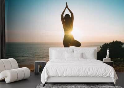 Woman practicing yoga and meditates on the mountain with ocean view at sunset. Young woman standing in vrksasana exercise, tree pose outdoor in the nature Wall mural