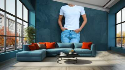 Young woman dressed in blank white t-shirt and blue jeans posing against the grey wall. Mockup for design print or t-shirt print store Wall mural