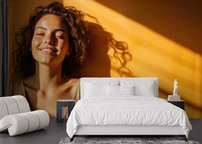 Young woman with curly hair smiling against an orange wall background with sunlight coming in from the side. Concept of joy and serenity. Wall mural