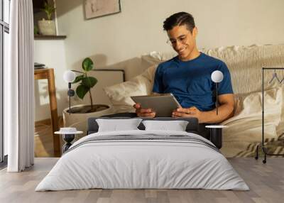 Attractive Hispanic man with glasses smiling wearing blue shirt siting on the sofa in the apartment using tablet  Wall mural