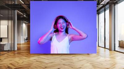 Young asian woman in white tank top wearing red headphone listen to music smiling on the purple neon light background. Wall mural