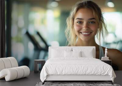 A cheerful woman is holding two dumbbells while smiling at the camera, enjoying her workout routine in a bright fitness center filled with equipment Wall mural