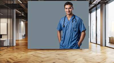 A male nurse with a beard, wearing blue scrubs and a stethoscope around his neck, stands with his hands in his pockets against a grey background. Wall mural