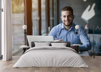 Young handsome man sitting in office with cup of coffee and working on project connected with modern cyber technologies. Businessman with notebook trying to keep deadline in digital marketing sphere. Wall mural