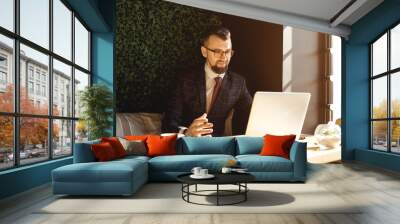 Young handsome man sitting in office with cup of coffee and working on project connected with modern cyber technologies. Businessman with notebook trying to keep deadline in digital marketing sphere. Wall mural