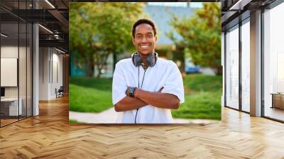 A young man smiles proudly with his arms crossed, wearing headphones, in a lively outdoor area Wall mural