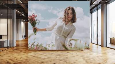 Upper body portrait of young woman in long white dress walking with a flower bouquet leaning to one side, against blue sky with clouds Wall mural