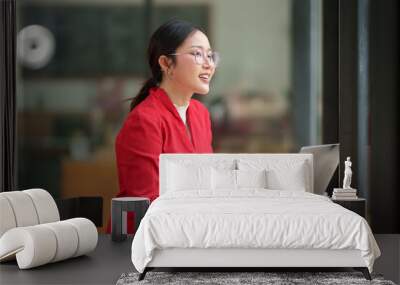 Confident Asian businesswoman in glasses sits on a chair in front of the counter, recording business financial goal plan online, analyzing, thinking, data entry on laptop of successful woman. Wall mural