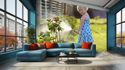 Cute toddler girl watering flowers from hose in summer garden Wall mural