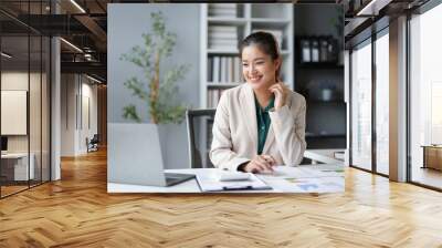 Asian businesswoman is smiling while working on a laptop and going over paperwork at her desk Wall mural