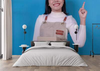 Smiling young asian woman in beige apron uniform holding package parcel box and pointing up with finger isolated on blue background Wall mural