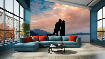 Silhouette of a cute couple in love at the moment of kissing with a background of a lake at sunset with clouds of warm tones illuminated by the sun. Wall mural