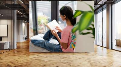 Girl sitting at hassock near window and reading book Wall mural