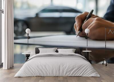 Close-up of a person’s hand signing a document on a table, with a car visible in the background. Wall mural