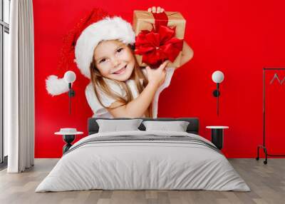 Happy smiling girl in Christmas cap with gift wrapped in craft paper and red ribbons on red background. Christmas. Happy family. Wall mural