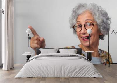 A joyful elderly woman with curly gray hair, wearing round glasses and a colorful floral cardigan, pointing with a playful smile, set against a clean white background Wall mural