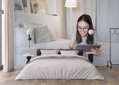 Cute teenager girl doing homework lying on bed at home. Young pretty girl wearing glasses writing down in notebook, studying online with tablet, distance learning, self education      Wall mural