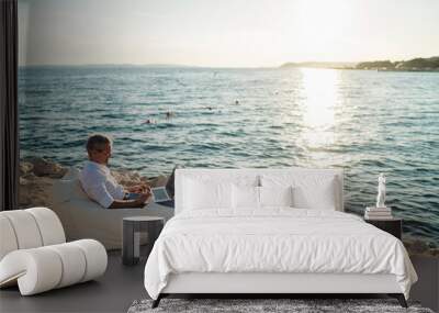 Senior man working on his laptop lying on deck chair on the beach during sunset Wall mural