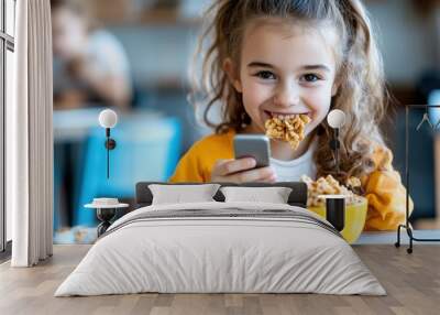 A cheerful young girl grins while eating a bowl of cereal, holding a smartphone, signifying modern lifestyle and the joy of playful morning routines. Wall mural