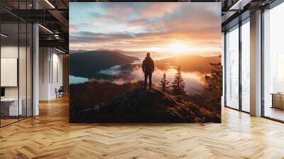 A lone person stands on a mountain peak overlooking a beautiful forested landscape with mist and warm colors of the sunset creating a serene and majestic view. Wall mural