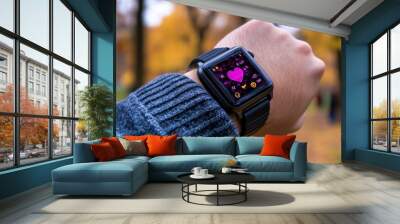 Close-up of a wrist wearing a modern smartwatch showing vibrant health and fitness data while outdoors in an autumn scene, emphasizing technology and lifestyle integration. Wall mural