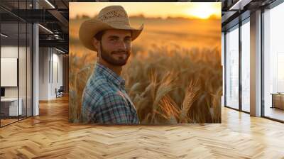 A man wearing a straw hat is standing in a field of tall golden wheat. He is looking up at the sun, which is setting in the background. Concept of peace and tranquility Wall mural