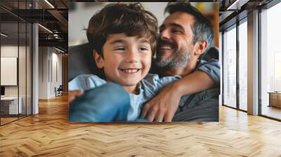 A father and his young son are captured sharing a joyful moment on a cozy sofa, both smiling and enjoying a precious bonding time in the warmly lit ambiance of their home. Wall mural