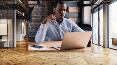 African male holding glasses while looking attentively at screen of laptop, analyzing important data Wall mural